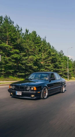 a black car driving down a street next to a forest, by Carlo Martini, pexels contest winner, baroque, bmw e 3 0, low quality photo, portrait shot 8 k, f11