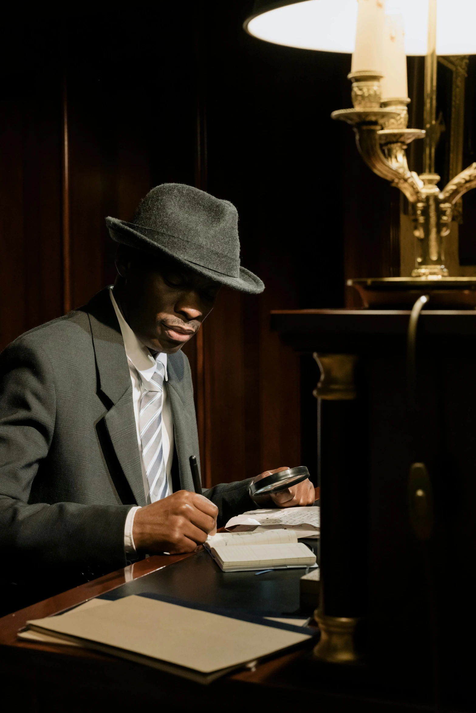 a man in a suit and hat sitting at a desk, by Clifford Ellis, unsplash, harlem renaissance, evening lighting, spying discretly, ( ( theatrical ) ), taken in the late 2010s