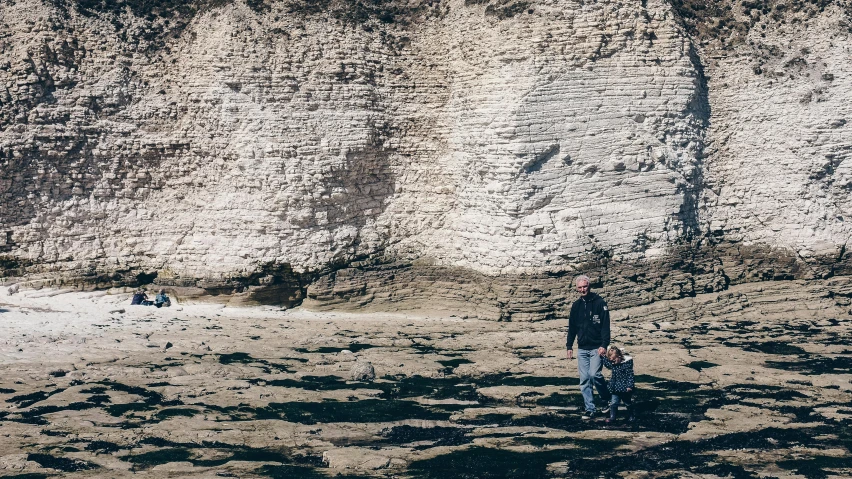 a man standing on top of a sandy beach next to a cliff, by Lee Loughridge, unsplash, les nabis, father with child, over a chalk cliff, texture, low quality photo