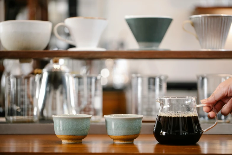 a person is pouring a cup of coffee, a still life, inspired by Miyagawa Chōshun, trending on unsplash, behind bar deck with bear mugs, dipped in polished blue ceramic, large jars on shelves, viridian and venetian red