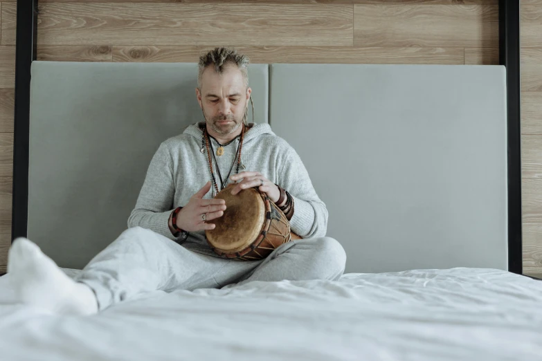 a man sitting on top of a bed holding a bag, an album cover, inspired by Peter Maxwell Ewart, pexels contest winner, hurufiyya, playing drums, holding a wood piece, contemplative, tribal yurta