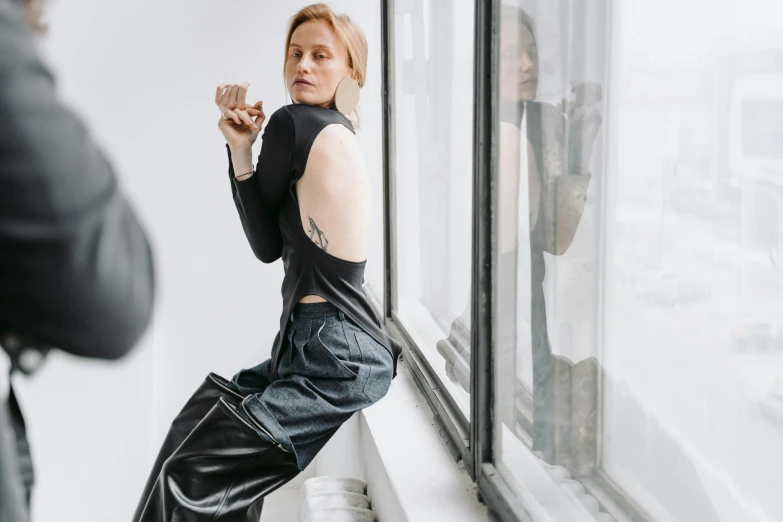 a woman standing in front of a window holding a cigarette, by Nina Hamnett, trending on pexels, bauhaus, black leather shiny jeans, evan rachel wood, artist wearing torn overalls, on a white table