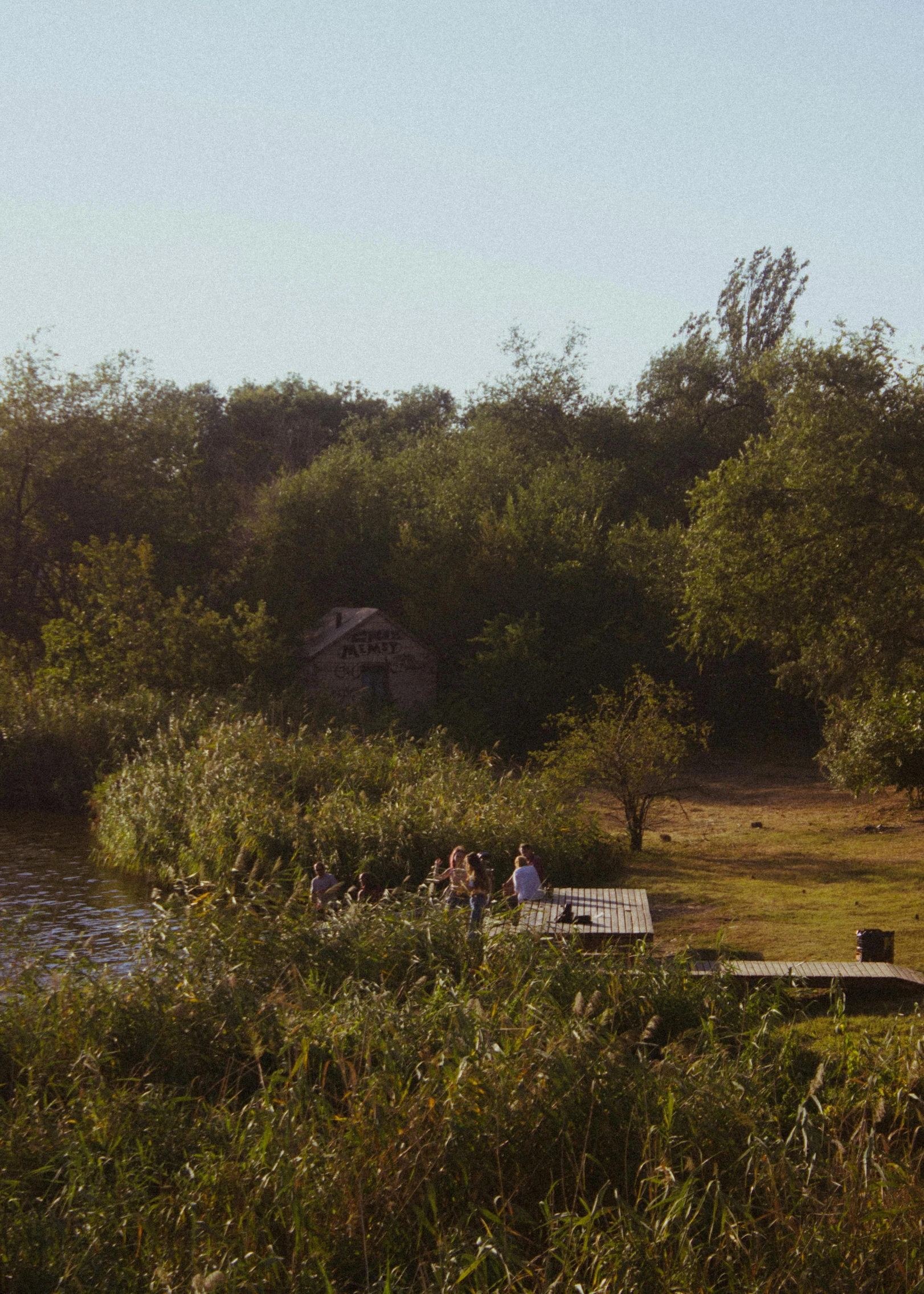 a group of people standing on top of a lush green field, small dock, woodland location, afternoon sun, wide film still