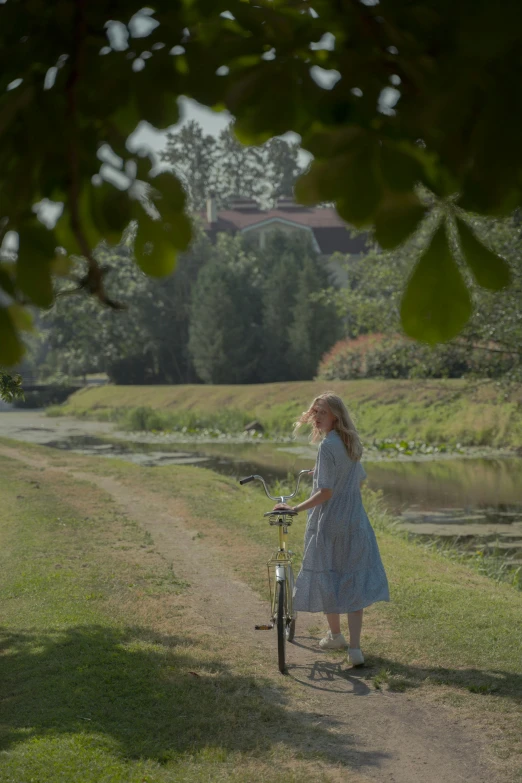 a woman riding a bike down a dirt road, inspired by Enrique Simonet, happening, imogen poots, near pond, ( ( theatrical ) ), cottagecore