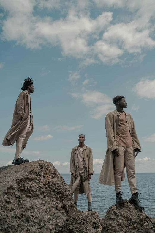 a group of men standing on top of a rock next to the ocean, an album cover, by Caro Niederer, unsplash, afrofuturism, brown robes, 3 actors on stage, ignant, ( ( theatrical ) )