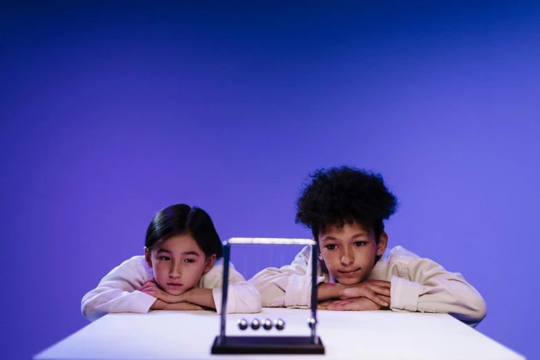 a couple of kids sitting at a table with a laptop, inspired by Storm Thorgerson, kinetic art, holding scale, purple scene lighting, close-up photograph, transparent