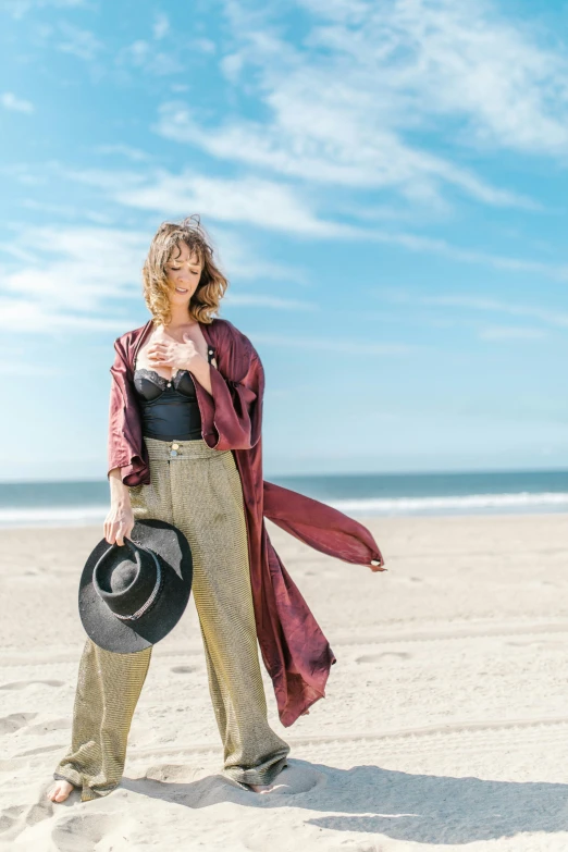 a woman standing on a beach holding a hat, a colorized photo, unsplash, renaissance, wearing a duster coat, santa monica beach, black and red silk clothing, wearing a linen shirt