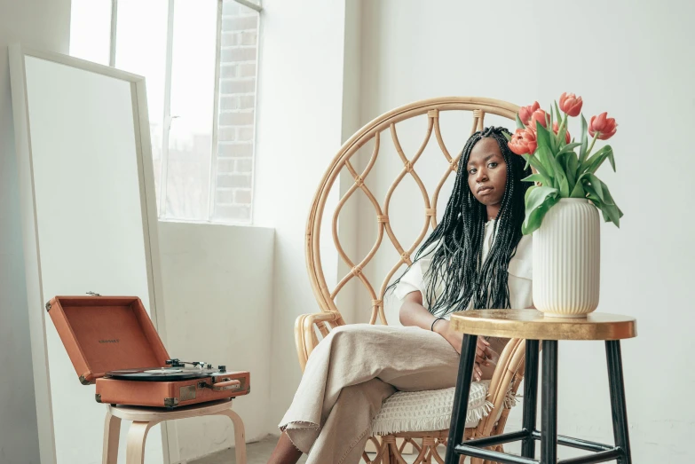 a woman sitting in a chair next to a vase of flowers, by Dulah Marie Evans, pexels contest winner, box braids, sitting on designer chair, calm afternoon, long dreadlocks