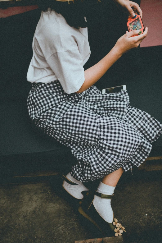 a woman sitting on a bench holding a cell phone, by Yasushi Sugiyama, trending on unsplash, plaid skirt, dark and white, wide skirts, studio photo