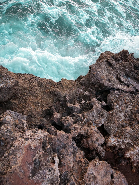 a man standing on top of a rocky cliff next to the ocean, by Daniel Lieske, pexels contest winner, lava texture, brown and cyan color scheme, high angle close up shot, waves and splashes