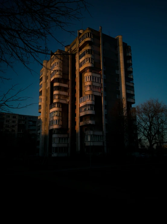 a tall building sitting in the middle of a park, an album cover, inspired by Elsa Bleda, unsplash, brutalism, soviet apartment building, dark moody backlighting, golden hour 8 k, soviet suburbs