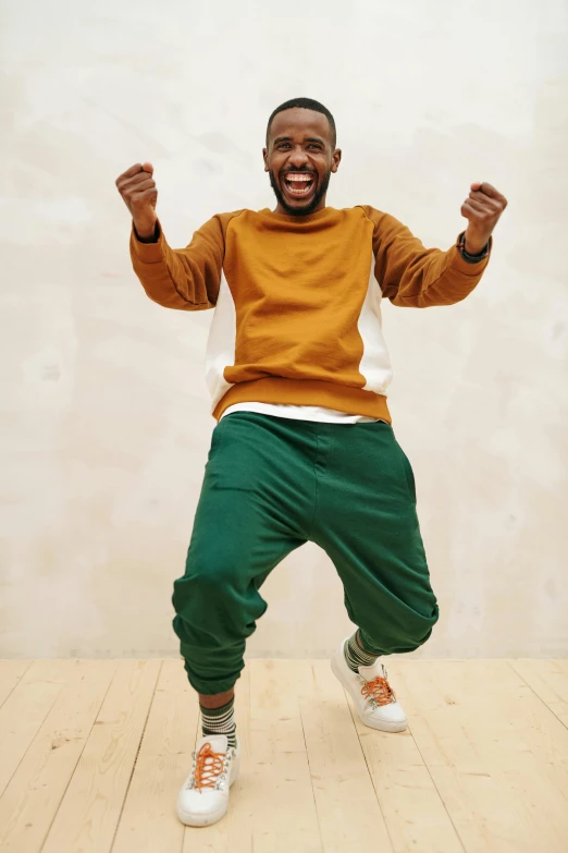 a man standing on a wooden floor with his arms in the air, inspired by Barthélemy Menn, trending on pexels, happening, wearing a green sweater, brown skin man with a giant grin, baggy pants, celebration