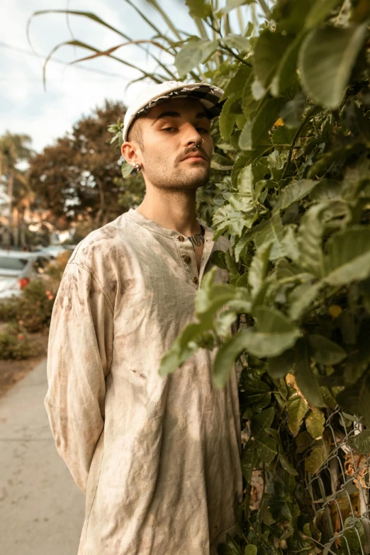 a man standing on a sidewalk next to a fence, an album cover, inspired by Elsa Bleda, renaissance, wearing ripped dirty flight suit, brown buzzcut, vine covered, lily frank