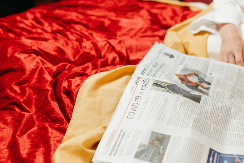 a person laying on a bed reading a newspaper, by Julia Pishtar, trending on unsplash, private press, red and gold cloth, wearing a velvet cloak, discovered for the first time, news coverage