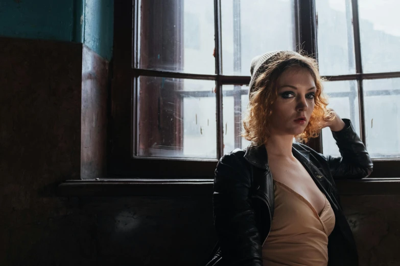 a woman sitting on a bench in front of a window, a portrait, inspired by Elsa Bleda, unsplash, renaissance, dressed in biker leather, natalie dormer, eighties-pinup style, standing in a dimly lit room