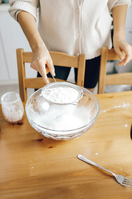 a woman mixing ingredients in a bowl on a wooden table, an album cover, inspired by Yukimasa Ida, trending on pexels, covered in white flour, glass tableware, -step 50, detailed product image