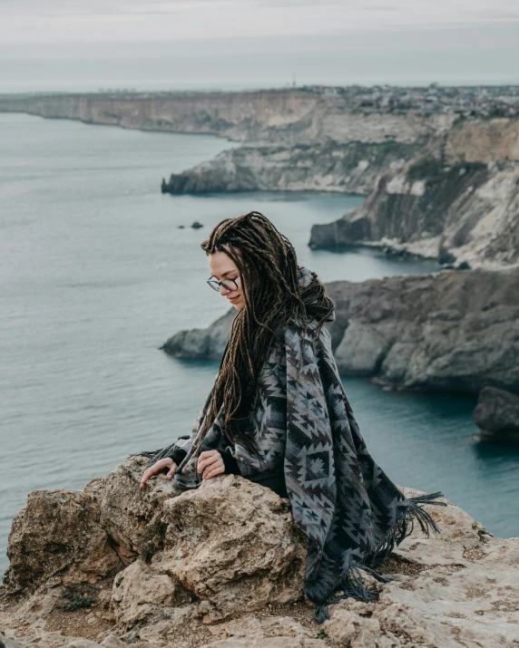 a woman sitting on top of a cliff next to the ocean, wrapped in a black scarf, 2019 trending photo