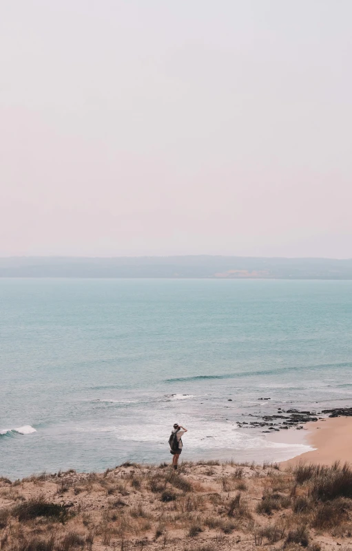 a man standing on top of a sandy beach next to the ocean, panoramic view of girl, unsplash photography, normandy, gif