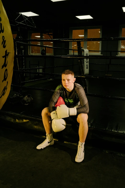 a man sitting in a boxing ring next to a punching bag, connor hibbs, confident pose, official photo, structure : kyle lambert