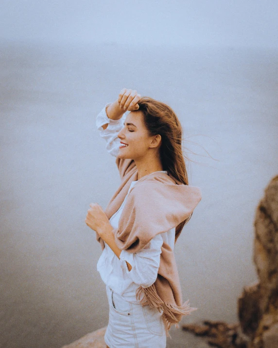 a woman standing on top of a rock next to a body of water, turning her head and smiling, white scarf, windy mane, muted color tones