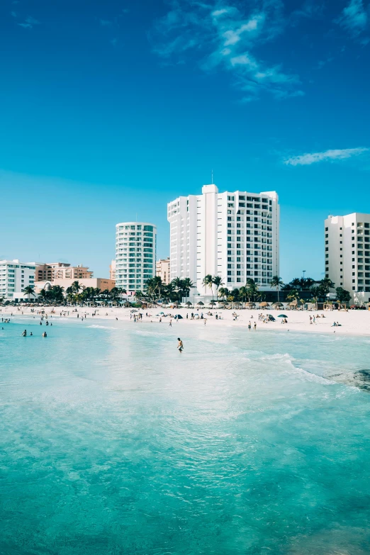 a group of people standing on top of a beach next to a body of water, white buildings, varadero beach, high rises, clear blue water