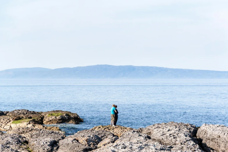 a man standing on top of a rock next to the ocean, by Alison Watt, people angling at the edge, cloudless-crear-sky, high-quality photo, hamar