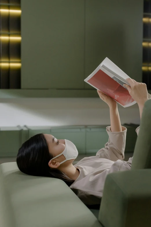 a woman laying on a couch reading a book, by Fei Danxu, unsplash contest winner, conceptual art, surgical mask covering mouth, futuristic product design, ignant, small chin