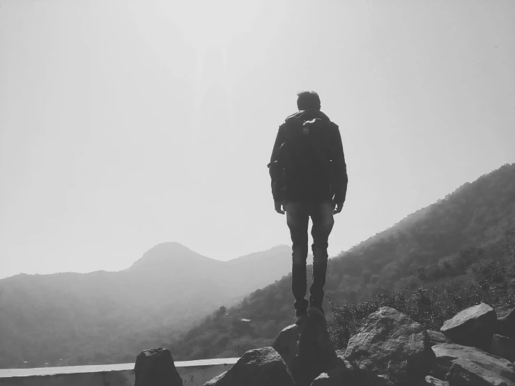 a man standing on top of a rock next to a body of water, a black and white photo, pexels, happening, background image, a man wearing a backpack, hills, profile pic