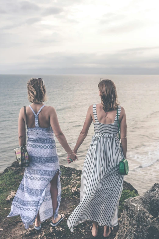 a couple of women standing on top of a cliff next to the ocean, trending on pexels, holding hands, lgbt, walking towards you, wearing dresses