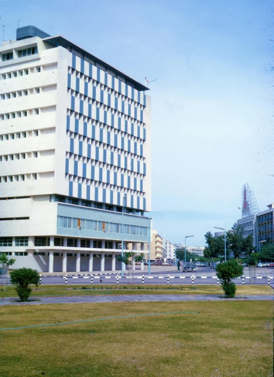 a large white building sitting on top of a lush green field, a colorized photo, brutalism, malaysian, city square, government archive photograph, street elevation