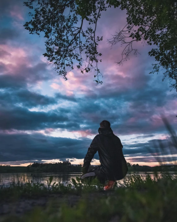 a person sitting next to a body of water under a cloudy sky, colorful sky, sitting under a tree, kneeling and looking up, instagram post