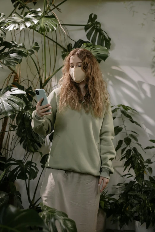 a woman wearing a face mask looking at her phone, inspired by Elsa Bleda, greenery, wearing an oversized sweater, posing for a picture, digital media