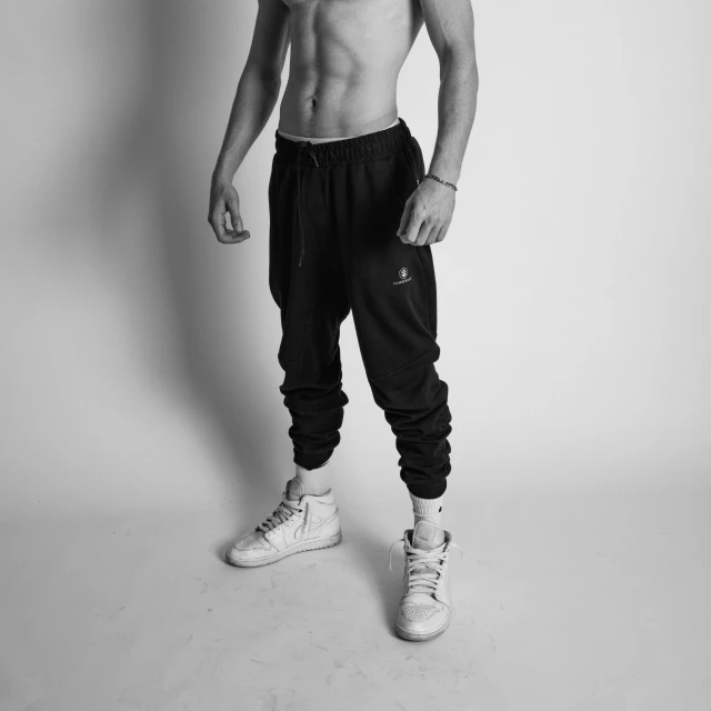 a shirtless man standing in front of a white wall, a black and white photo, unsplash, renaissance, wearing a tracksuit, zirconium pants, black clothing, product render
