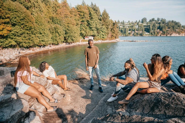 a group of people sitting on top of a rock next to a body of water, by Jessie Algie, pexels contest winner, man is with black skin, vancouver, lush surroundings, childish gambino