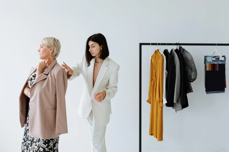 two women standing next to each other in front of a rack of clothes, by Emma Andijewska, trending on pexels, minimalism, dressed in an old white coat, reaching out to each other, people at work, presenting wares