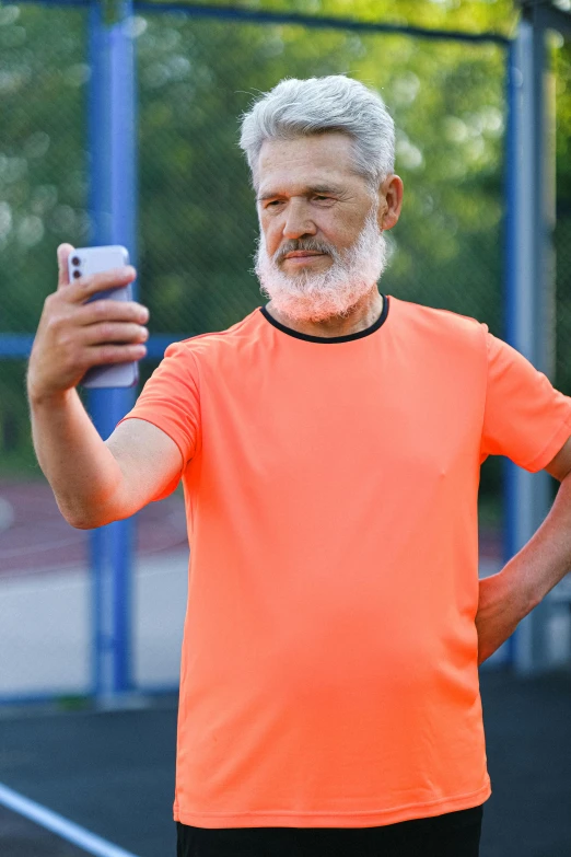 a man standing on a tennis court holding a cell phone, a picture, shutterstock, happening, long grey beard, wearing an orange t shirt, hgh, wearing fitness gear