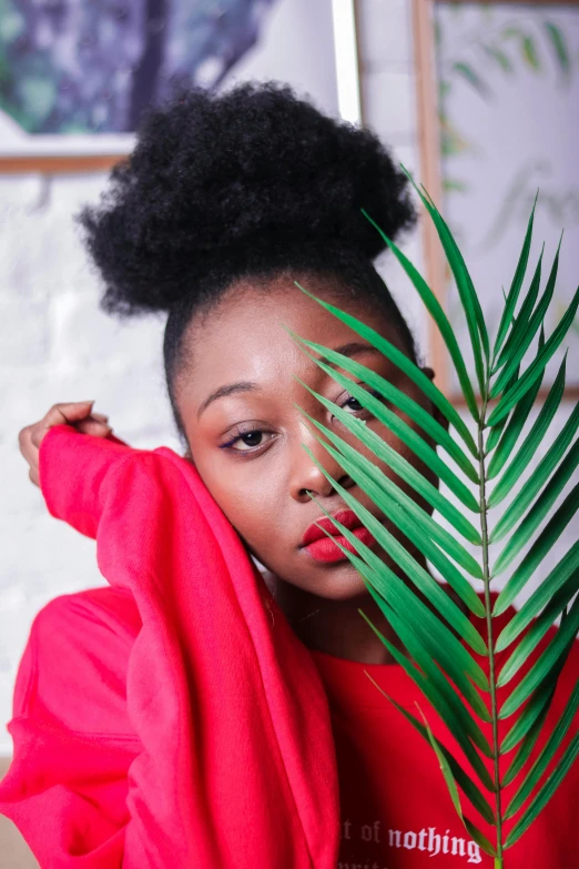 a woman holding a palm leaf in front of her face, an album cover, trending on pexels, afrofuturism, wearing a red turtleneck sweater, girl with messy bun hairstyle, at a fashion shoot, black teenage girl