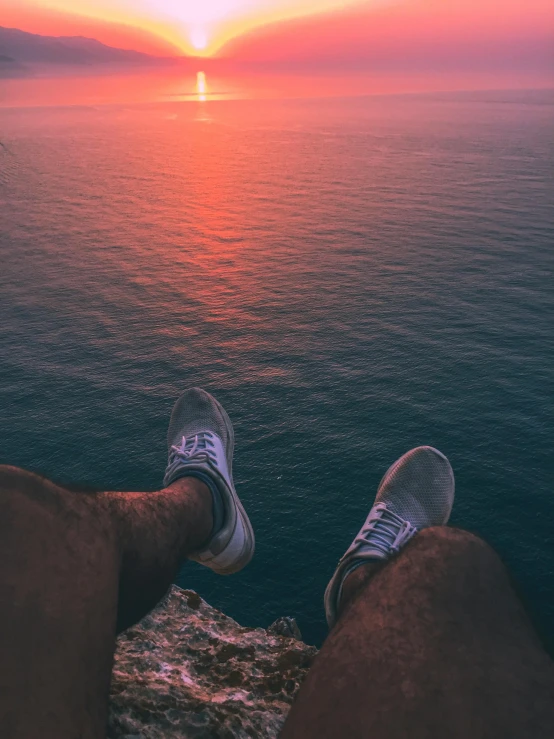 a person sitting on top of a rock next to the ocean, sneaker photo, ((sunset)), towering high up over your view, 8k octan photo