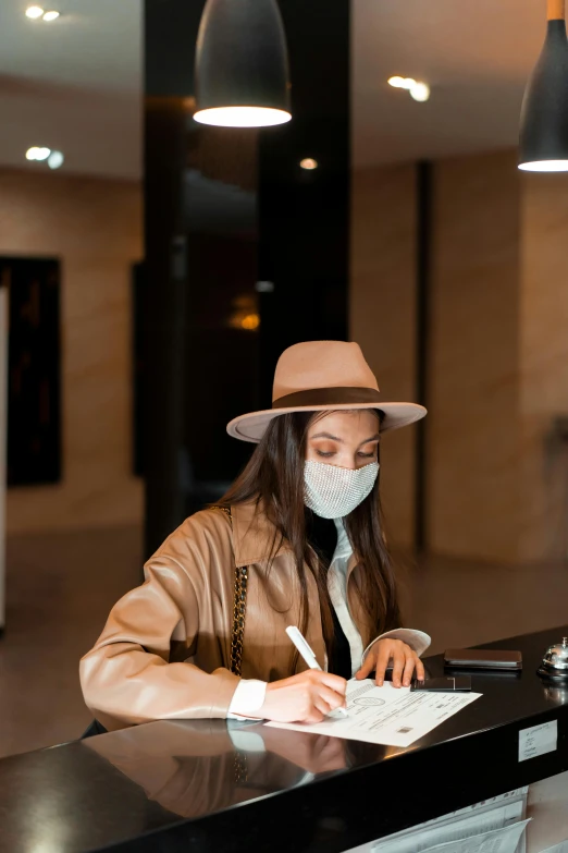 a woman sitting at a desk with a mask on, pexels contest winner, in a hotel hallway, caracter with brown hat, signing a bill, wearing a brown leather coat