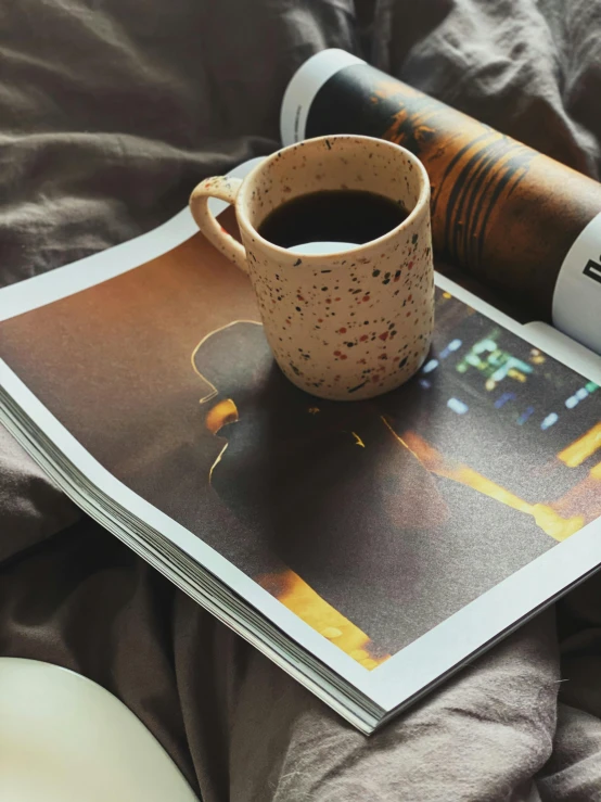 a magazine sitting on top of a bed next to a cup of coffee, pexels contest winner, photorealism, grainy poster art, brown and cream color scheme, dusk setting, portait photo