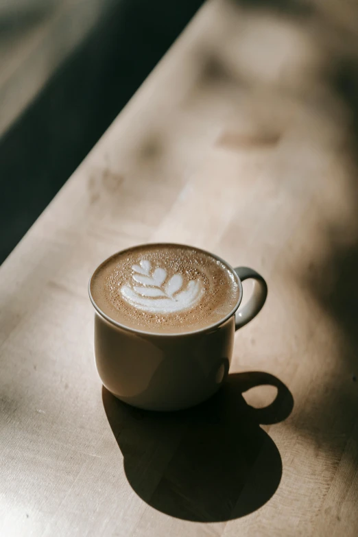 a cup of coffee sitting on top of a wooden table, jen atkin, latte art, bathed in light, ignant