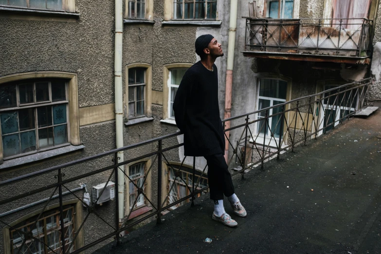 a man standing on top of a balcony next to a building, an album cover, by Attila Meszlenyi, pexels contest winner, casual black clothing, moscow, modest, androgynous person