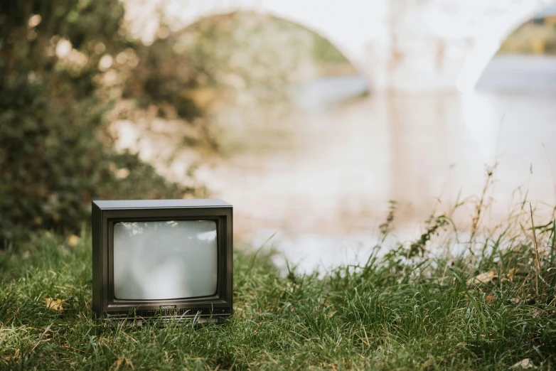 a small television sitting on top of a lush green field, a cartoon, unsplash, video art, bokeh on side of the river, 1 9 8 0 s photo, cast, urban surroundings