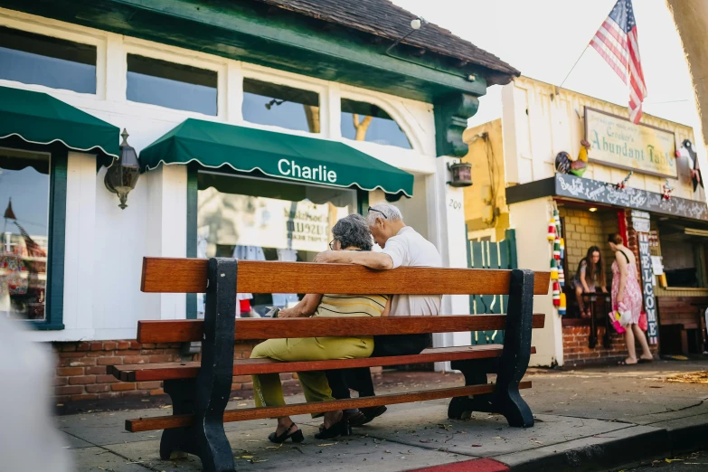 a couple of people that are sitting on a bench, a photo, by Charles Martin, restaurant, comforting and familiar, charlie, neighborhood