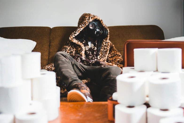 a person sitting on a couch surrounded by rolls of toilet paper, an album cover, unsplash, visual art, ski mask, brown clothes, cinematic outfit photo, gas mask