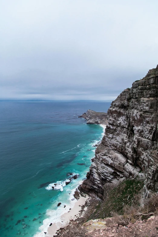 a man standing on top of a cliff next to the ocean, by Peter Churcher, pexels contest winner, south african coast, teal color graded, “ aerial view of a mountain, big overcast