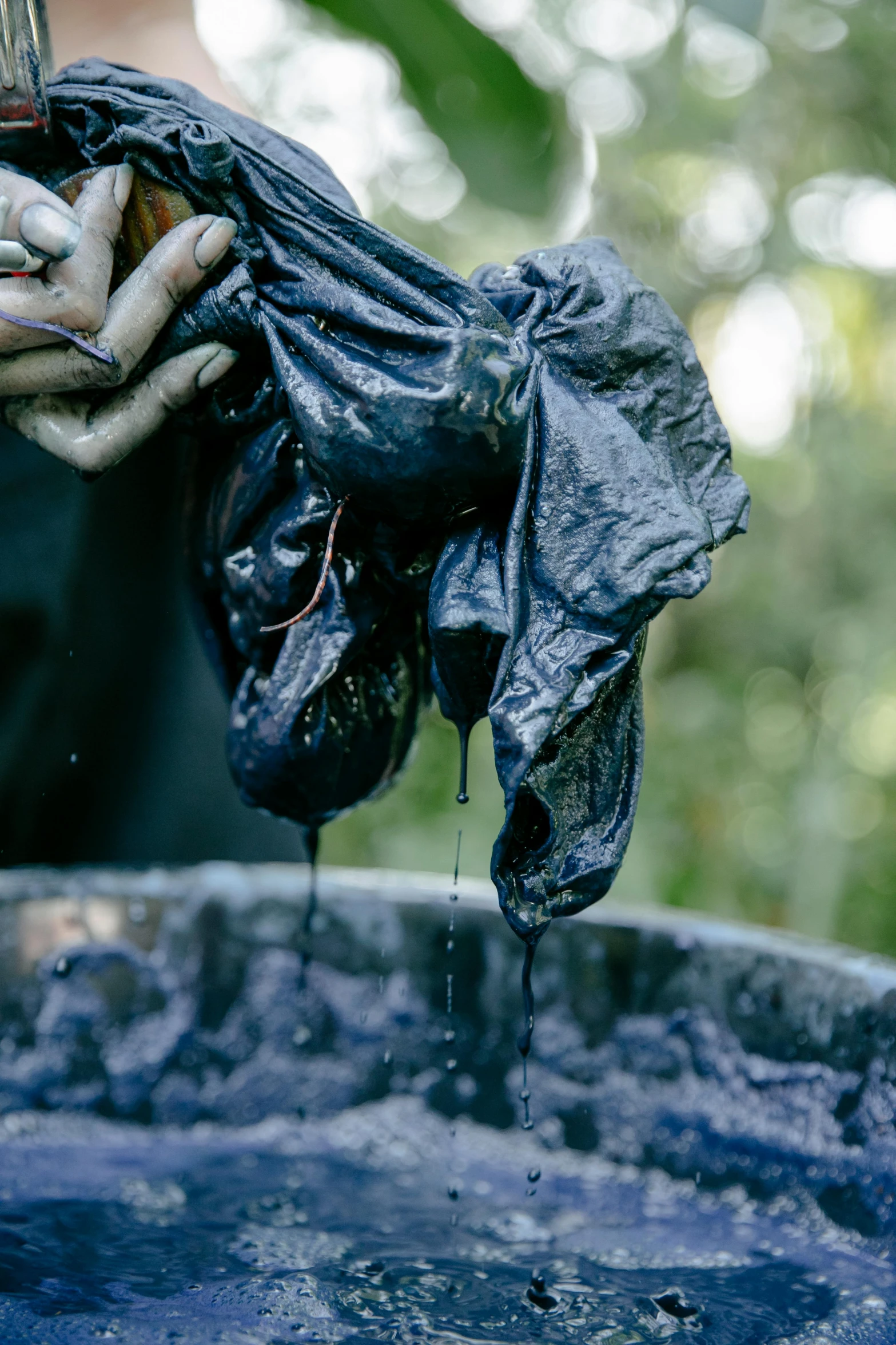 a person that is holding something in a bucket, covered with black goo, indigo, worn clothes, lush surroundings