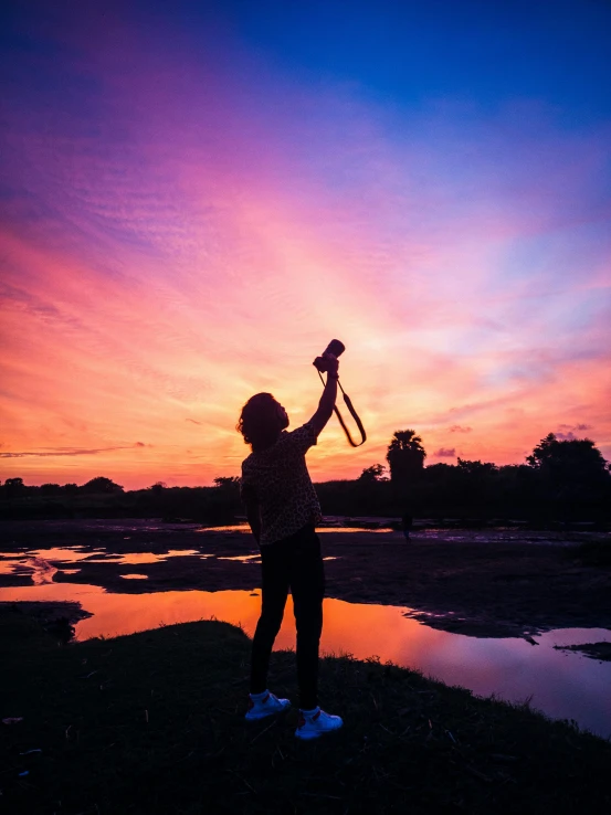 a woman holding a camera up in the air, pexels contest winner, violet and yellow sunset, holding flask in hand, profile pic, ( land )