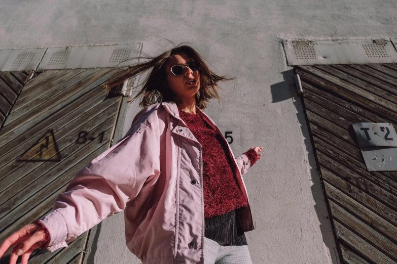 a woman standing in front of a building, by Olivia Peguero, pexels contest winner, wearing an aviator jacket, ((pink)), playful pose, faded colours