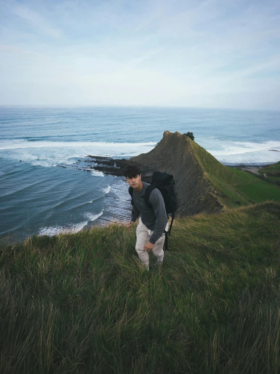 a man standing on top of a hill next to the ocean, a man wearing a backpack, in a large grassy green field, trecking, ocean swells